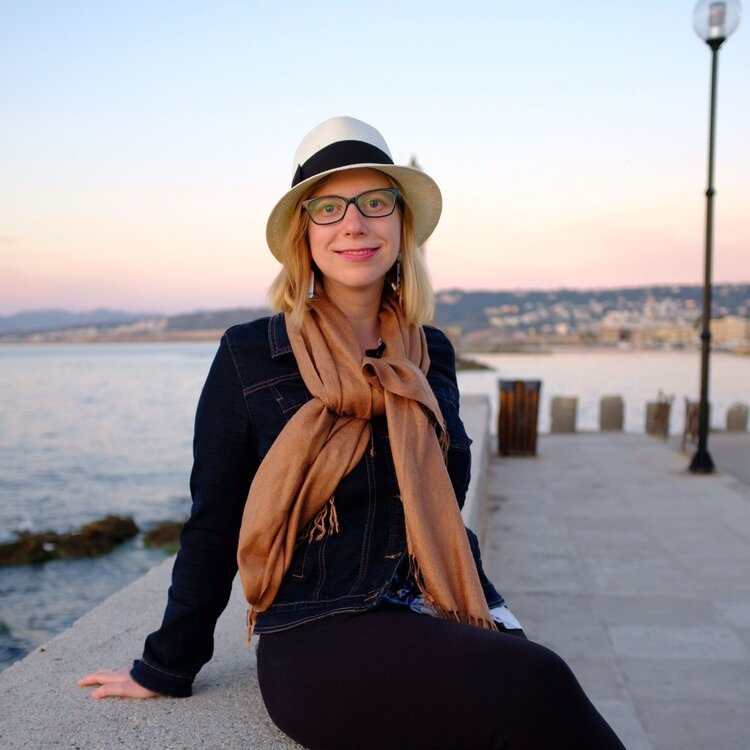 Helen sits besides the ocean at sunset. She wears a wide brimmed hat and a brown scarf, and smiled at the camera.