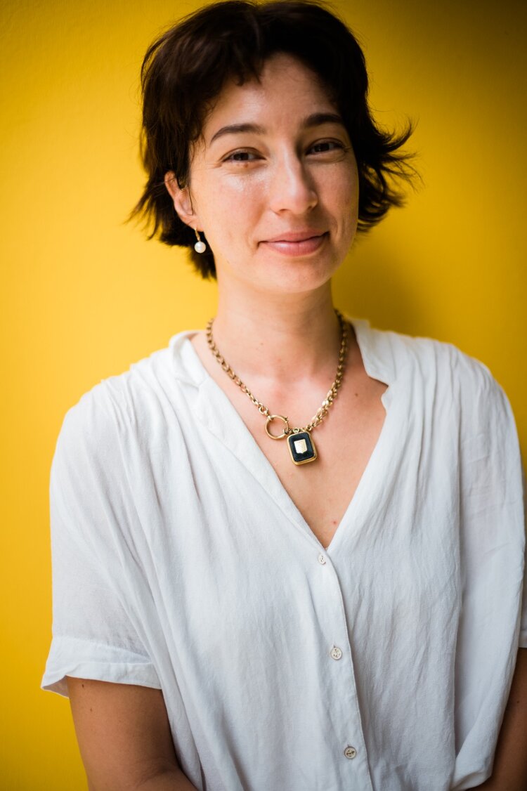 Johanna wears a white button up shirt and gold necklace. She smiles at the camera against a yellow wall.
