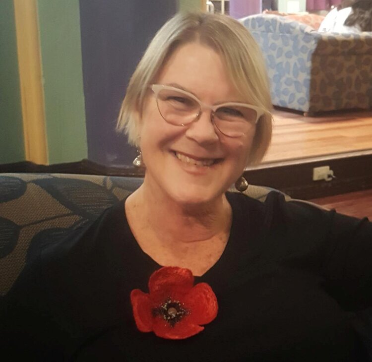 Natalie wears glasses and smiles at the camera. She wears a black shirt and a poppy pin.