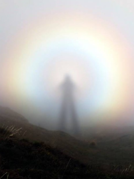 A shadow or figure of a person behind a raimbow circle of light, standing on a mountain.