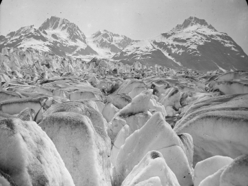 A black and white photo of chunks of glacier ice, with snow-covered mountains in the background.